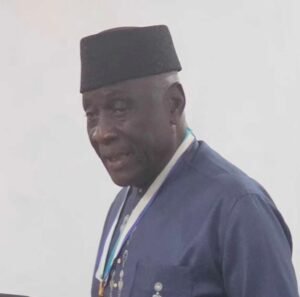 | The Calabash Newspaper Man in a dark blue traditional outfit with a black cap and white lanyard, looking to the side.