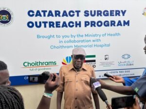 Elderly man in a tan shirt and cap speaks to reporters in front of a banner that reads 'Cataract Surgery Outreach Program.' Multinational logos and microphones surround him.
