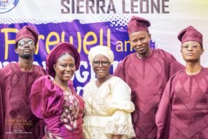 Five people in maroon traditional attire pose for a group photo at a farewell event with a purple banner in the background.—informational
