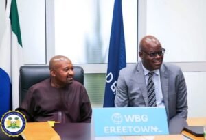 | The Calabash Newspaper Two men sit at a conference table in a meeting room, smiling; one wears a maroon shirt, the other a gray suit, with a blue WBG Ereetown sign and flags in the background.