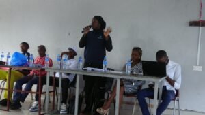| The Calabash Newspaper A woman standing and speaking into a microphone at a long table in a panel setup, while several people sit and listen with water bottles in front of them.