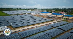 Aerial view of a large solar farm with rows of panels, blue-roofed buildings, and workers in safety vests nearby.