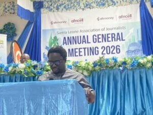 | The Calabash Newspaper Man speaking at a blue podium during the Sierra Leone Association of Journalists Annual General Meeting 2026, with blue floral decor and a large banner in the background.