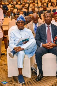 Front-row conference attendees: a man in light blue traditional attire with a head wrap and headset reads documents beside a man in a dark suit, as others watch from the audience.