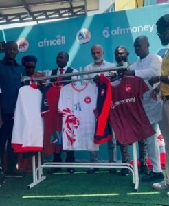 Group of men at a sporting event standing behind a rack of colorful football jerseys (red, white, and maroon) displayed outdoors on a turf surface, with a blue sponsor backdrop showing Africell and Afrimoney logos.
