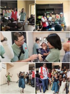 | The Calabash Newspaper Volunteers in green shirts and a red vest engaging with children at a classroom event, raising hands in greeting or activity moment.