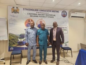 | The Calabash Newspaper Three men pose indoors in front of a banner for a Stakeholder Consultative Meeting on the National Railway Masterplan and feasibility studies, with a train image depicted on the banner.