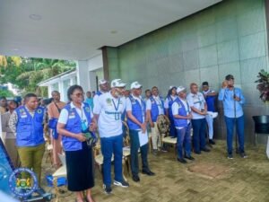 Group of census workers in blue vests standing together at an outdoor event, some holding papers and wearing lanyards as a crowd watches.