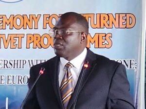 | The Calabash Newspaper Man in a dark suit and striped tie speaks at a podium with two red-lit microphones, in front of a banner.