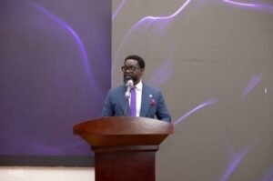 | The Calabash Newspaper Man in a blue suit and purple tie speaks at a wooden podium on a stage with purple-lit backdrop.