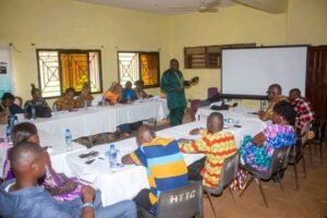 | The Calabash Newspaper Group of people seated around long tables in a workshop, a speaker stands at the front near a projector screen in a yellow-walled room.