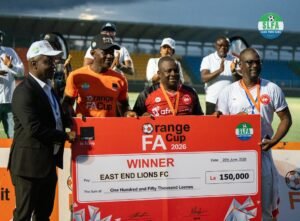 Three men hold a large red prize check reading WINNER East End Lions FC at a football awards ceremony in a stadium