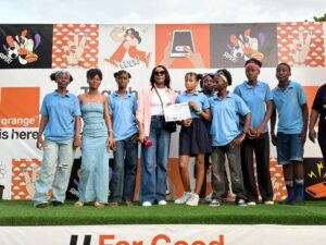 Group of youths in blue polo shirts with a woman at center holding a certificate, posing for a photo on a decorated stage during a community event.