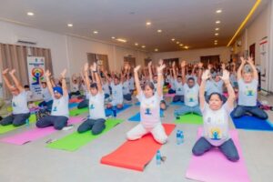 Group of people in a large bright hall practicing yoga on colorful mats, arms raised overhead in a synchronized pose; participants wear matching white shirts and water bottles are visible nearby.