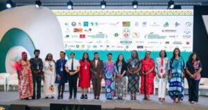 | The Calabash Newspaper Diverse group of 14 professionals on a conference stage, wearing lanyards, posing for a photo with a banner of logos behind them.