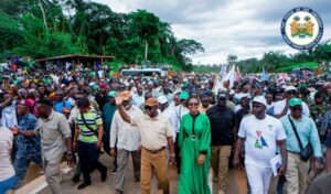 Large crowd gathered on a dirt road for a public event, with officials walking at the front and a government seal in the top-right corner.