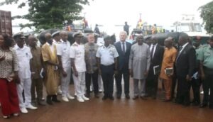 Group of officials in uniform and formal attire posing for a photo outdoors, with vehicles and security personnel in the background at an official event