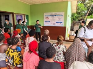 Community health workers in green shirts give a health education talk to a seated crowd of men and women outside a green building, with posters on the wall.