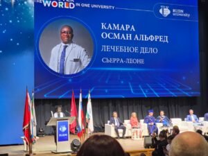 Conference stage at a university event with a large blue banner showing a doctor portrait and Cyrillic text; panelists sit on white chairs, flags lined up in front.