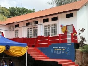 Exterior of a school with a red staircase leading to a blue platform labeled 'Albert Academy Science Lab', white walls, and an orange roof; yellow and blue bunting and balloons decorate the area.