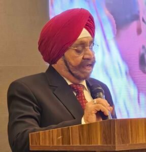 | The Calabash Newspaper Man wearing a red turban and glasses, speaking into a microphone at a wooden podium on a stage.