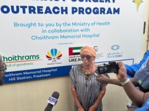 | The Calabash Newspaper Woman in a striped dress and orange headscarf being interviewed by reporters in front of a banner for a health outreach program, with logos and microphones visible in the background, including SLBC/Noor Dubai branding and UAE flag elements.