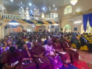Large hall full of people in purple and brown robes attending a formal ceremony, with a balcony and chandeliers above and gold drapery along the railings.