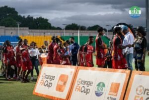 Youth soccer players in red jerseys line up on a sunny stadium field as officials hand out trophies near orange FA Cup banners.