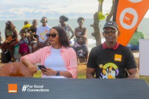 Two people sit at a table on a beach during a community event, with a crowd behind them and a banner that reads 'For Good Connections.'