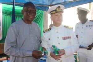 Two men at a formal ceremony swap green ribbons; a civilian in gray traditional attire stands with a naval officer in white dress uniform under a blue canopy.