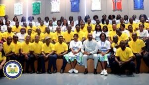 | The Calabash Newspaper Group photo of students and adults in a hall, many wearing yellow shirts, posing for a ceremony with a crest on the floor left corner and colorful shirts on the wall behind.
