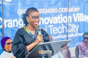 Woman in a black gown speaks into a microphone at a podium during a ceremony, with a blue banner in the background reading 'Innovation Village Tech City'.