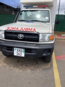 Front view of a white Toyota ambulance with red'AMBULANCE' lettering on the hood, blue and red emergency lights, and license plate AXG 163.