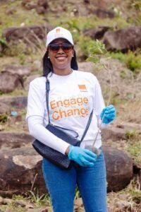 Woman planting a sapling outdoors, wearing a white 'Engage for Change' shirt and blue gloves.