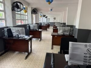 Empty computer lab with rows of desks and computer workstations, fans on the walls, and blue and yellow balloons overhead.