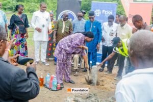 Group of people at a groundbreaking ceremony; a woman in purple digs into the soil while others look on.