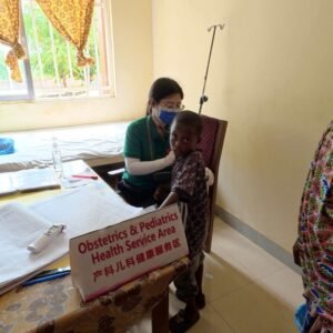 | The Calabash Newspaper Healthcare worker in a green shirt and blue mask checks a young boy in a clinic; sign reads Obstetrics & Pediatrics Health Service Area.