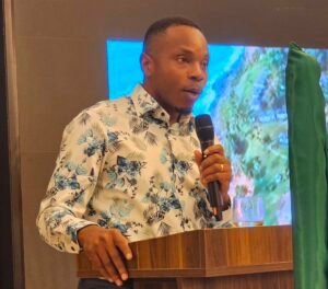 | The Calabash Newspaper Man in a tropical-pattern shirt speaks into a handheld microphone at a wooden podium during a presentation.