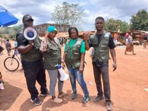 | The Calabash Newspaper Four volunteers in green vests stand together at an outdoor market; one man holds a megaphone and speaks to passersby.