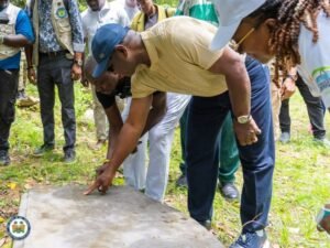 | The Calabash Newspaper Group of people outdoors bending toward a concrete slab as a man in a blue cap points at it while others watch in a park-like setting.