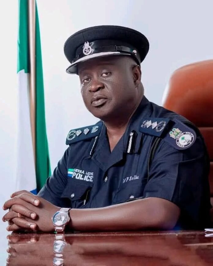 Portrait of a Sierra Leone police officer in uniform seated at a desk with folded hands, flag in the background.