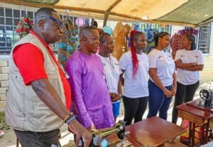 Instructor in red shirt and beige vest demonstrates sewing to a group of students under a canopy, with sewing machines on a table behind them.