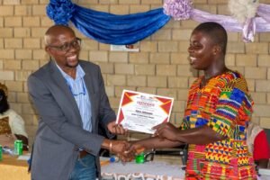 Two people shake hands as a certificate is presented during a ceremony; the man in a suit smiles while the recipient wears a colorful dress in front of a brick wall decorated with blue and purple fabric.