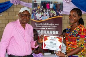 | The Calabash Newspaper Older man in a pink shirt hands a certificate to a young woman in a colorful dress during a community awards ceremony, with a Kush Rehabilitation and Empowerment Project banner in the background.
