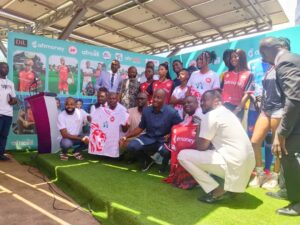 | The Calabash Newspaper Group of people posing on a green stage at a sports event, some wearing red and white jerseys, with sponsor banners in the background.