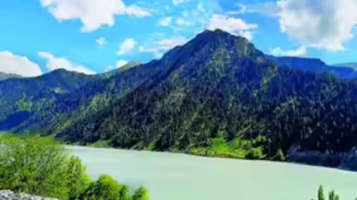 Mountain lake with turquoise-green water, forested mountains, and a bright blue sky with scattered clouds in the background.