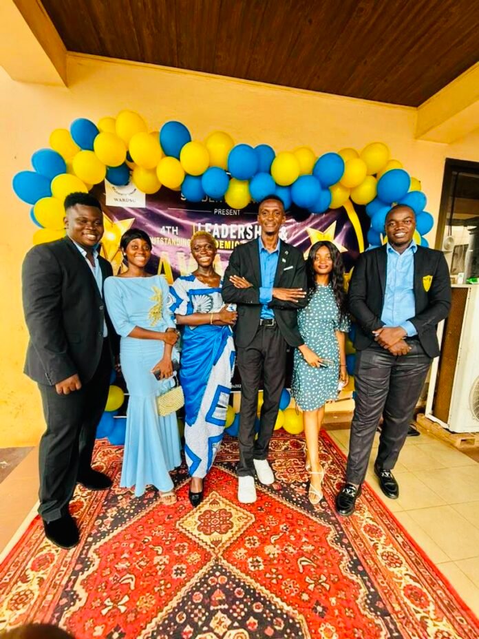 Group of six well-dressed young adults posing under a yellow-blue balloon arch at a formal awards event with a decorative rug and banner behind them