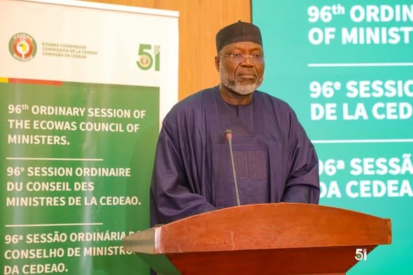 Man in a dark purple traditional outfit speaks at a wooden podium during the ECOWAS Ministers' 96nd Ordinary Session, with banners in the background.