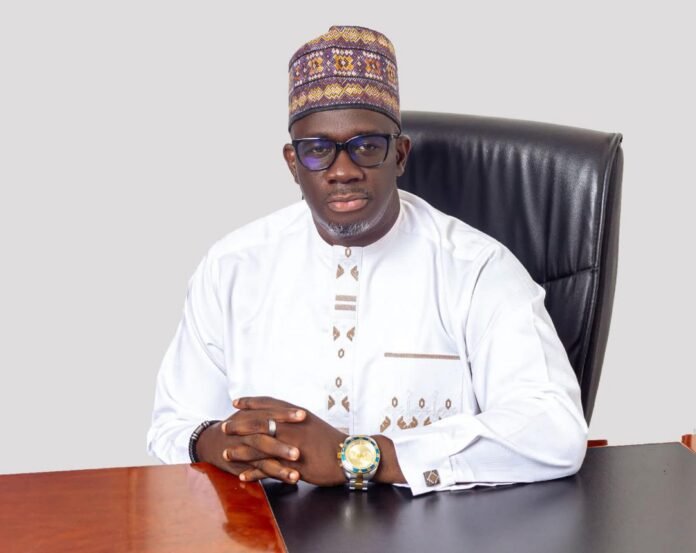 Portrait of a man in a white traditional outfit and patterned cap, seated at a desk with folded hands and a gold watch.
