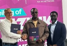 Africell Signs Drizilik as Brand Ambassador to Inspire the Next Generation Three men in a ceremony shake hands; two hold navy folders with the africell logo against a bright magenta backdrop.